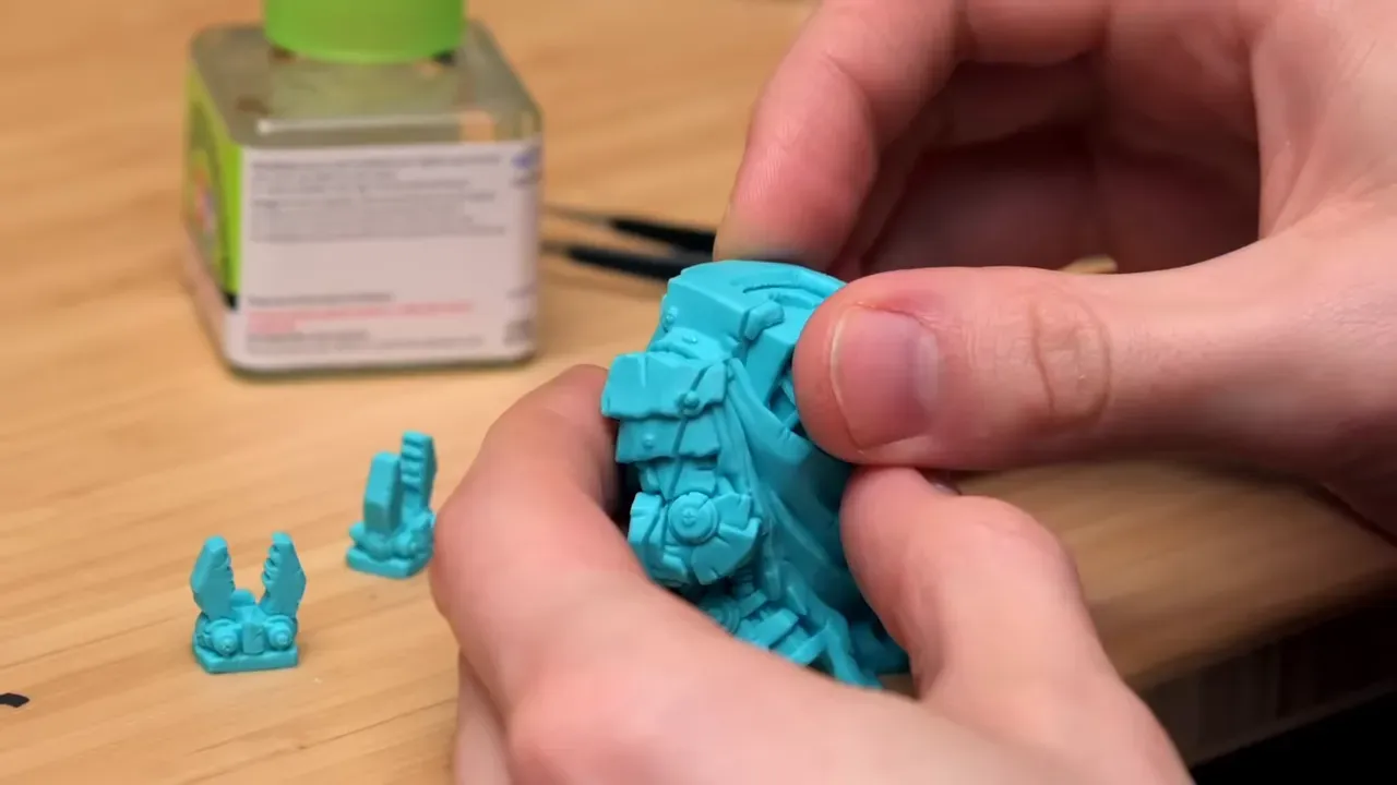 Deux mains assemblant les deux moitiés turquoise d une grande miniature avec un flacon vert à côté